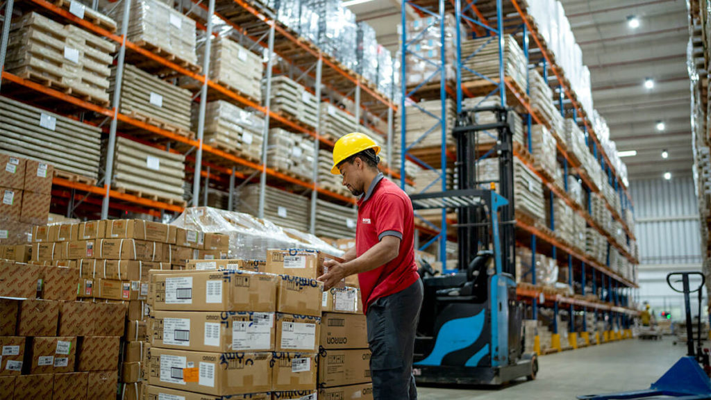 Trabalhador de logística com capacete e uniforme confere caixas em um grande centro de distribuição da Favorita com prateleiras altas repletas de mercadorias. A cena representa a organização e eficiência necessárias na logística para e-commerce, com foco no armazenamento, separação e expedição de pedidos.