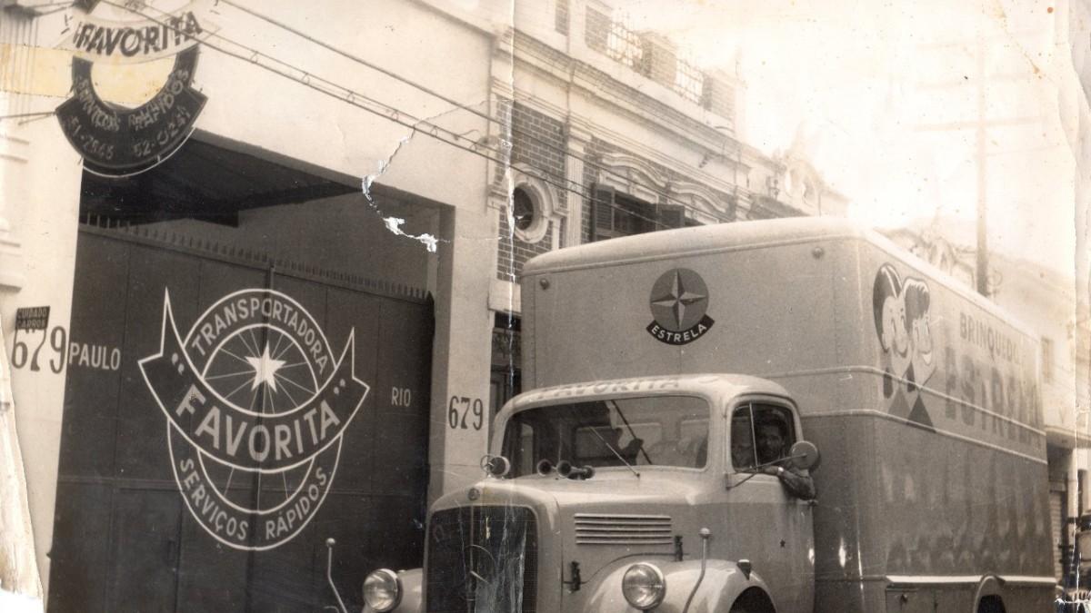 Imagem histórica que retrata a história da Favorita Transportes, com caminhão antigo em frente à uma de suas primeiras sedes, simbolizando o início das operações logísticas no Brasil.